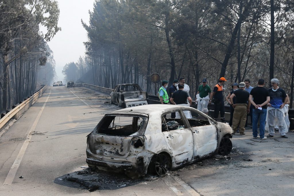 Bomberos, agentes de policía y Protección Civil, y personal forense hacen un alto en el trabajo junto a dos de los vehículos quie ardieron cuando las llamas envolvieron de golpe la carretera.